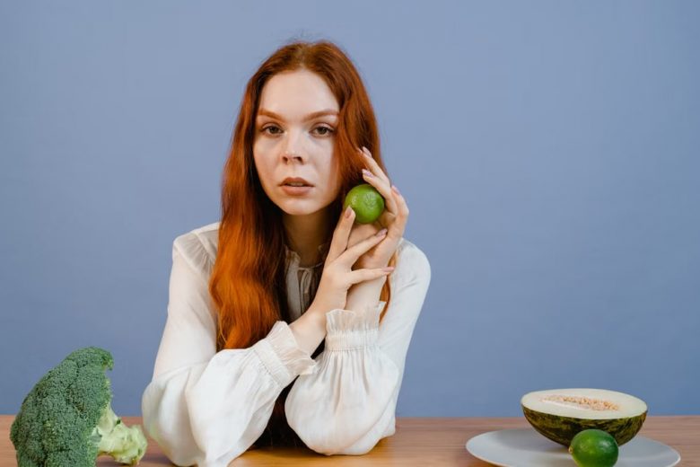Woman with red hair holding lime surrounded by fresh produce on table.